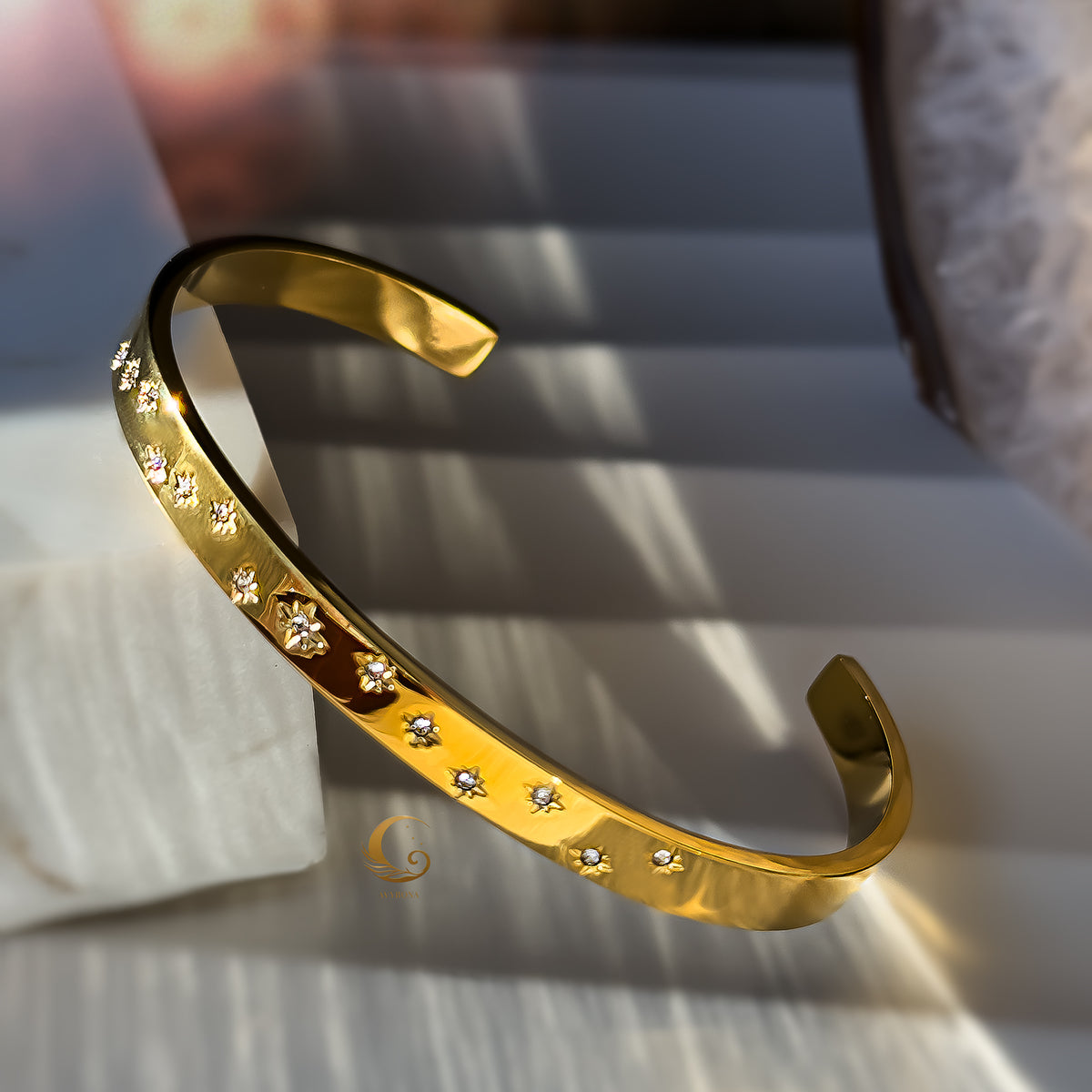 Single golden cuff bracelet with delicate engraved stars and stones, styled on a soft white stone.