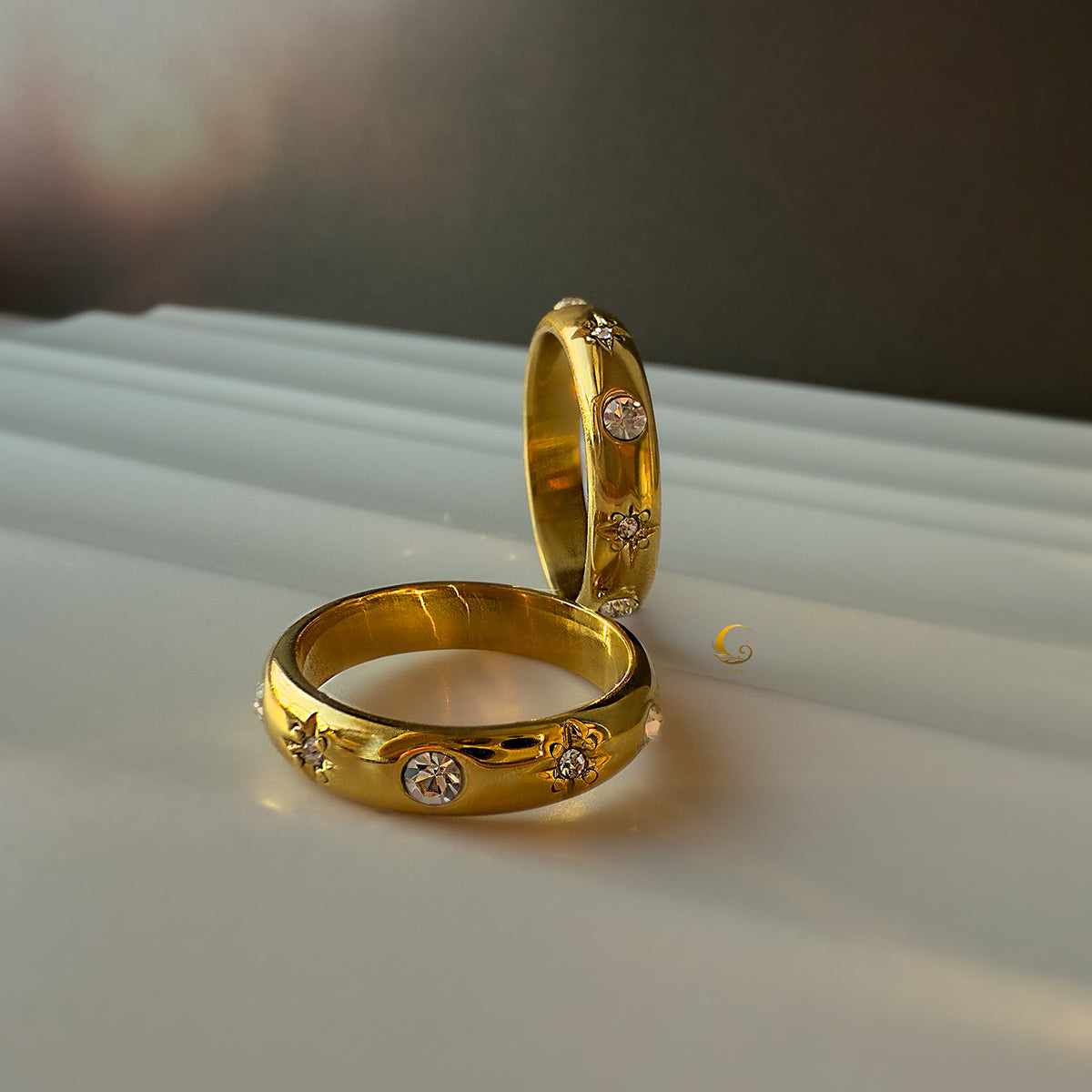 Close-up of two gold Celestial Glow rings with star engravings and crystals on white background
