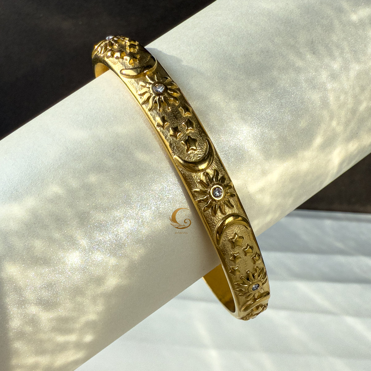  Side view of a gold-plated open bangle bracelet featuring engraved cosmic symbols—sun, crescent moons, and stars—on a soft rolled background with shimmering light.