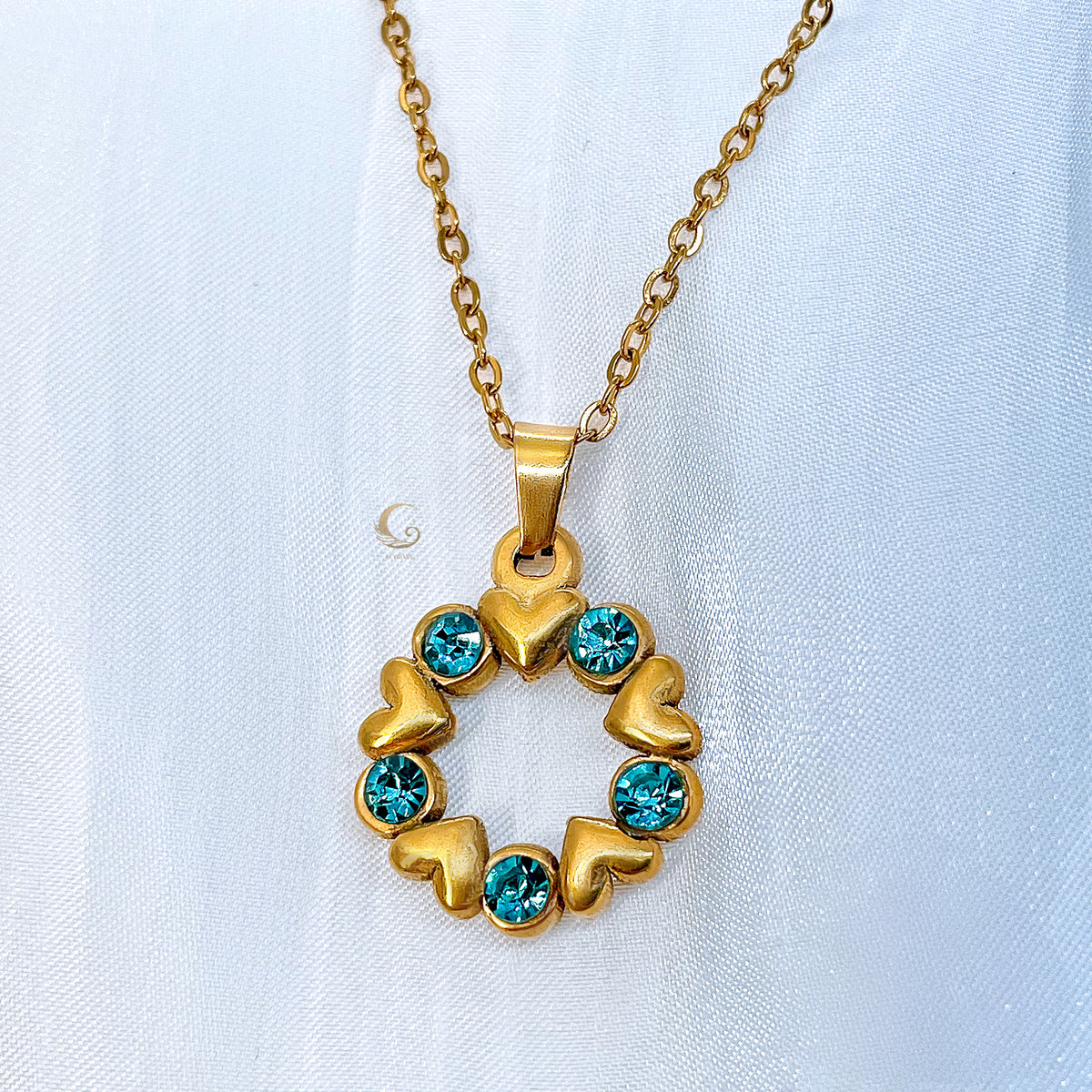 Close-up of the Elina necklace with round golden pendant featuring six hearts and six blue crystals in a circle, on a medium-thick gold chain.
