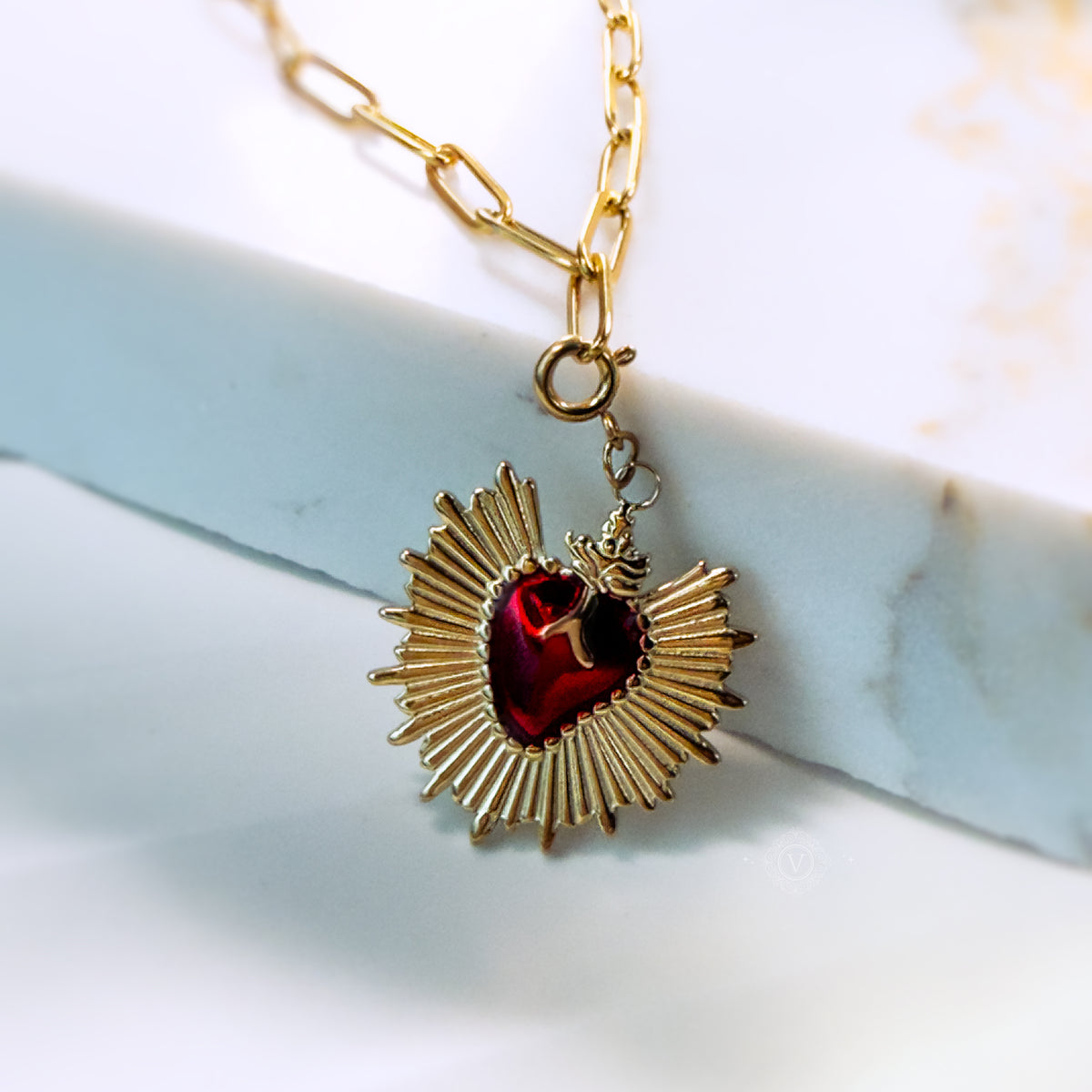 Eloria Sacred Heart necklace displayed on a white marble surface, showcasing the fiery red enamel and sunburst gold rays.