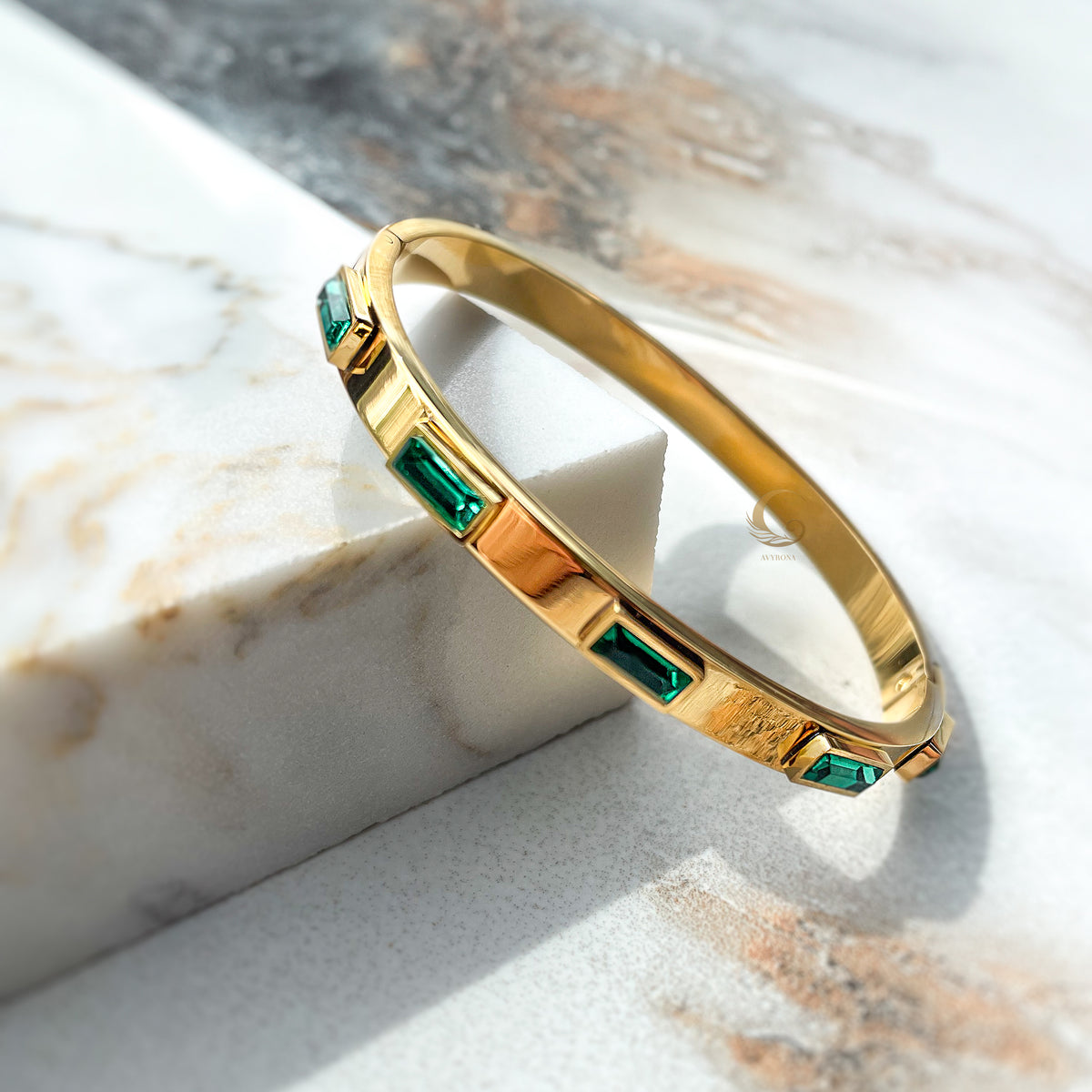 Gold-plated bangle bracelet with emerald green rectangular crystals, displayed on a marble surface in angled light.