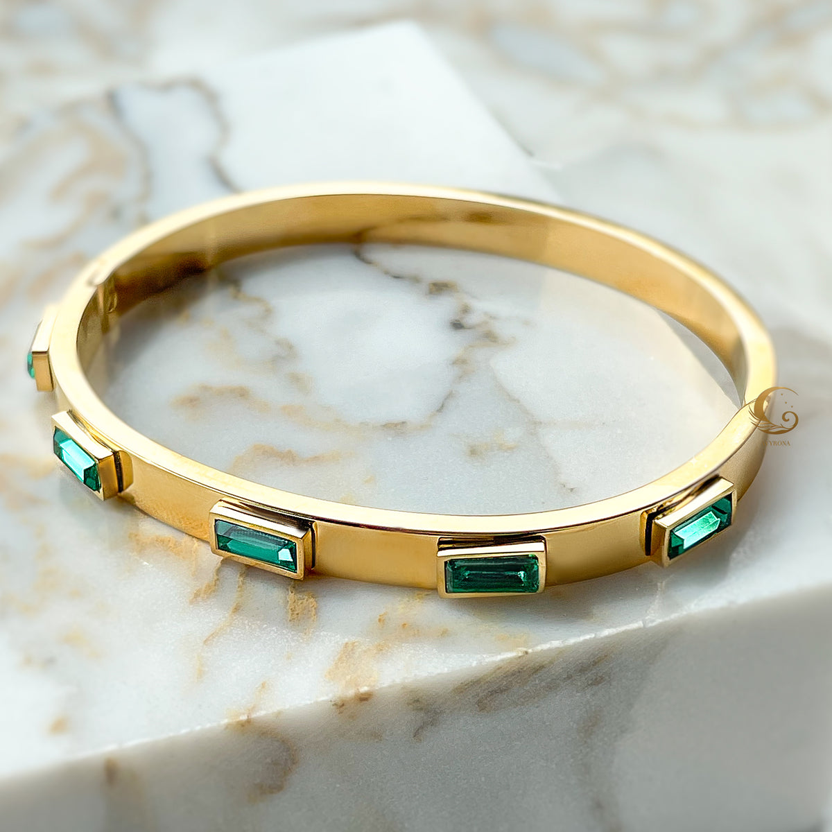 Top view of gold-plated stainless steel bangle bracelet with green crystals, photographed on a marble background.