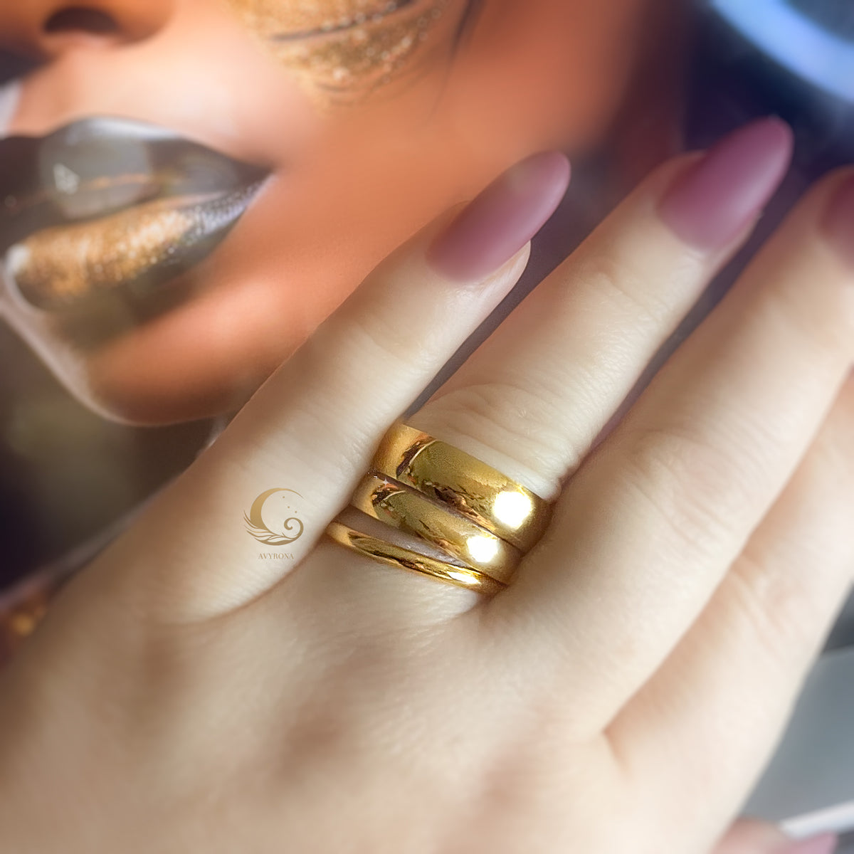 Hand wearing three stacked gold rings of different widths, styled with warm-toned makeup.
