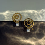 Gold Thaleia earrings with pearl-like drops on black and gold surface, glowing in contrast