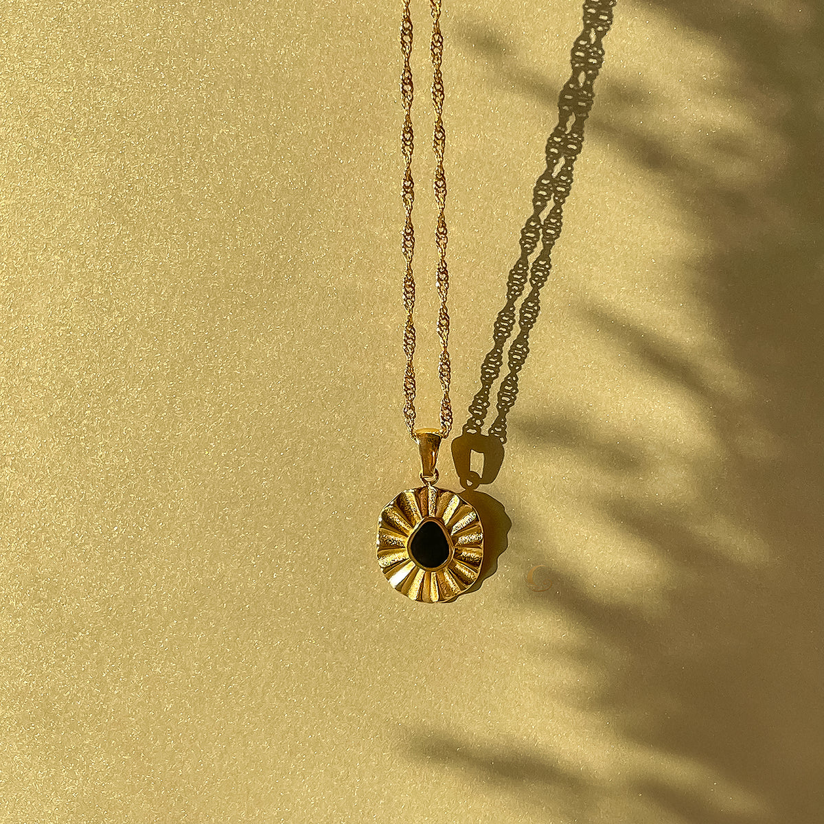 Liore Black Flower Necklace on a shimmering golden background with soft shadows. The floral pendant is styled with a twisted gold chain, creating a warm, sunlit effect.