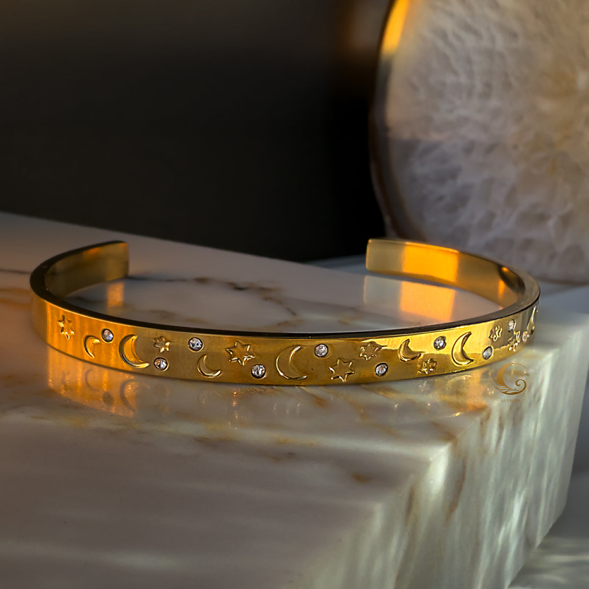 Gold cuff bracelet with engraved moons, stars, and clear zircons, displayed on marble with soft golden light.