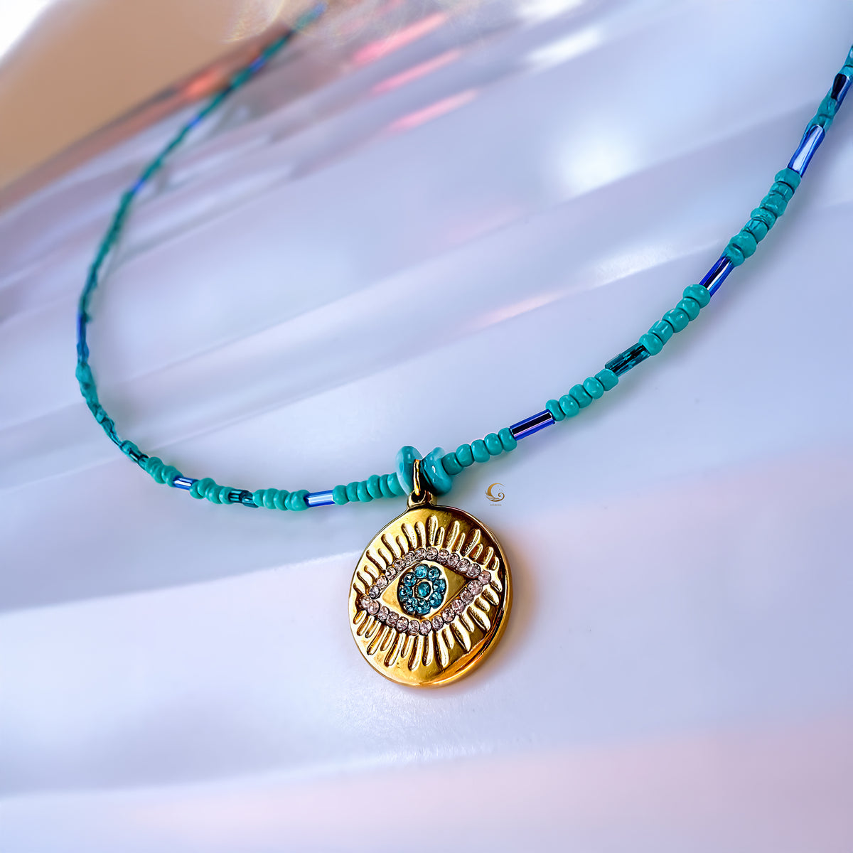 Marisel necklace with turquoise beads and gold Evil Eye charm photographed on a soft pearlescent surface.