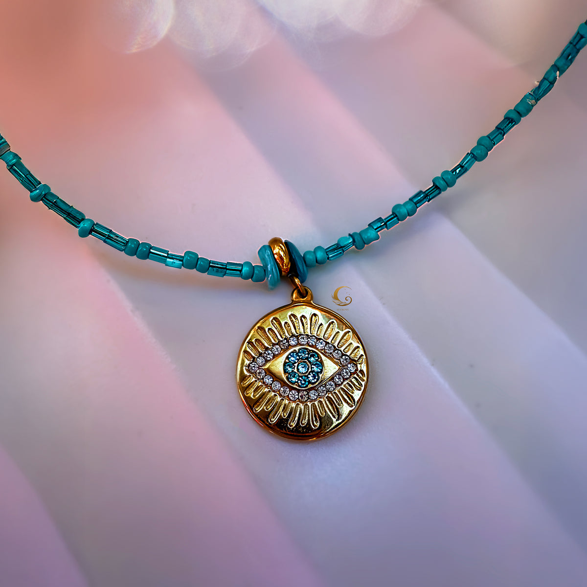 Turquoise beaded Marisel Evil Eye necklace on satin background, showing reflective gold pendant and soft light tones.