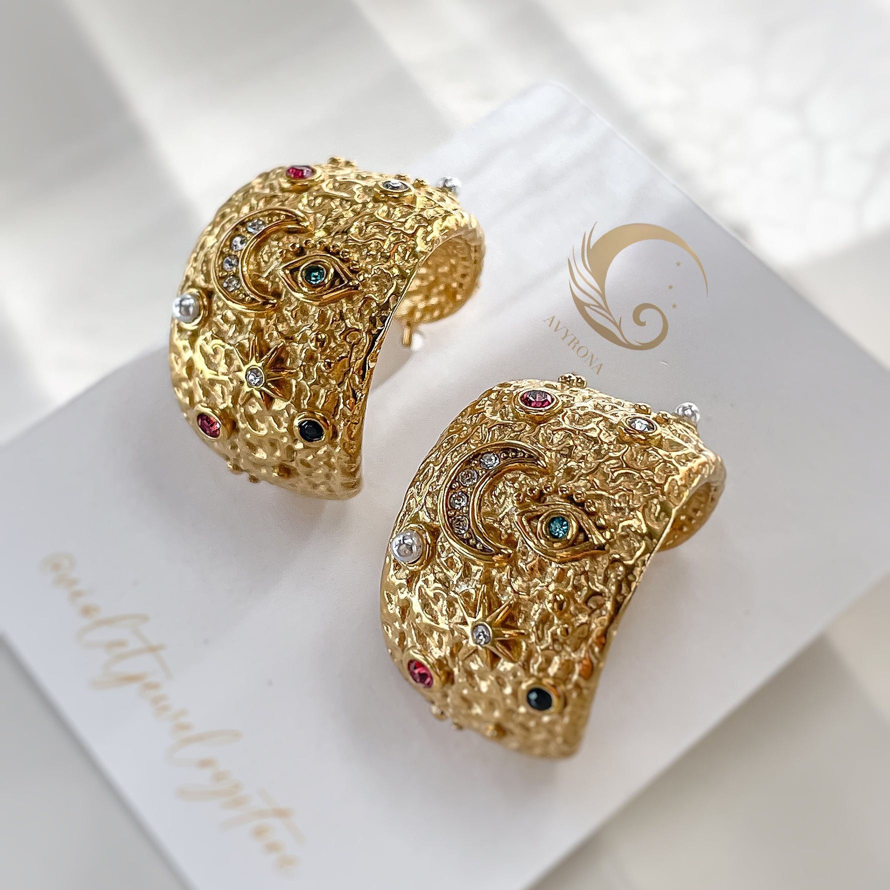 Celestial hoop earrings laid on branded Avyrona card, showing cosmic design with moon, eye, and star symbols in colorful accents.