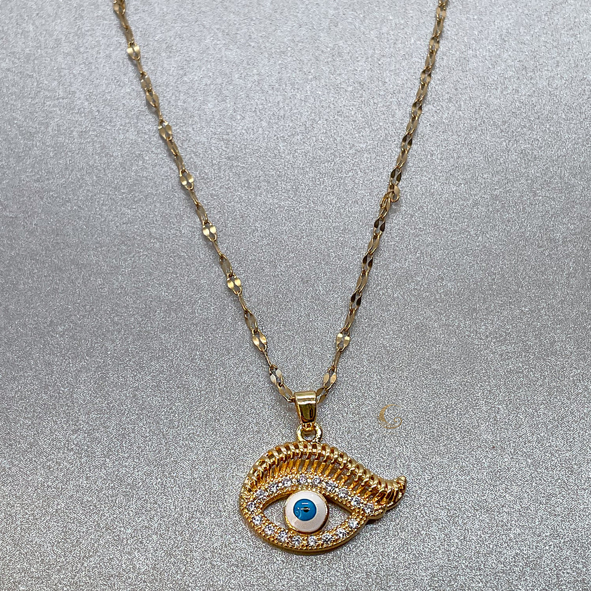 Gold evil eye necklace pendant close-up, twisted chain and cubic zirconia detail on silver textured background, symbolic protection jewelry, Avyrona

