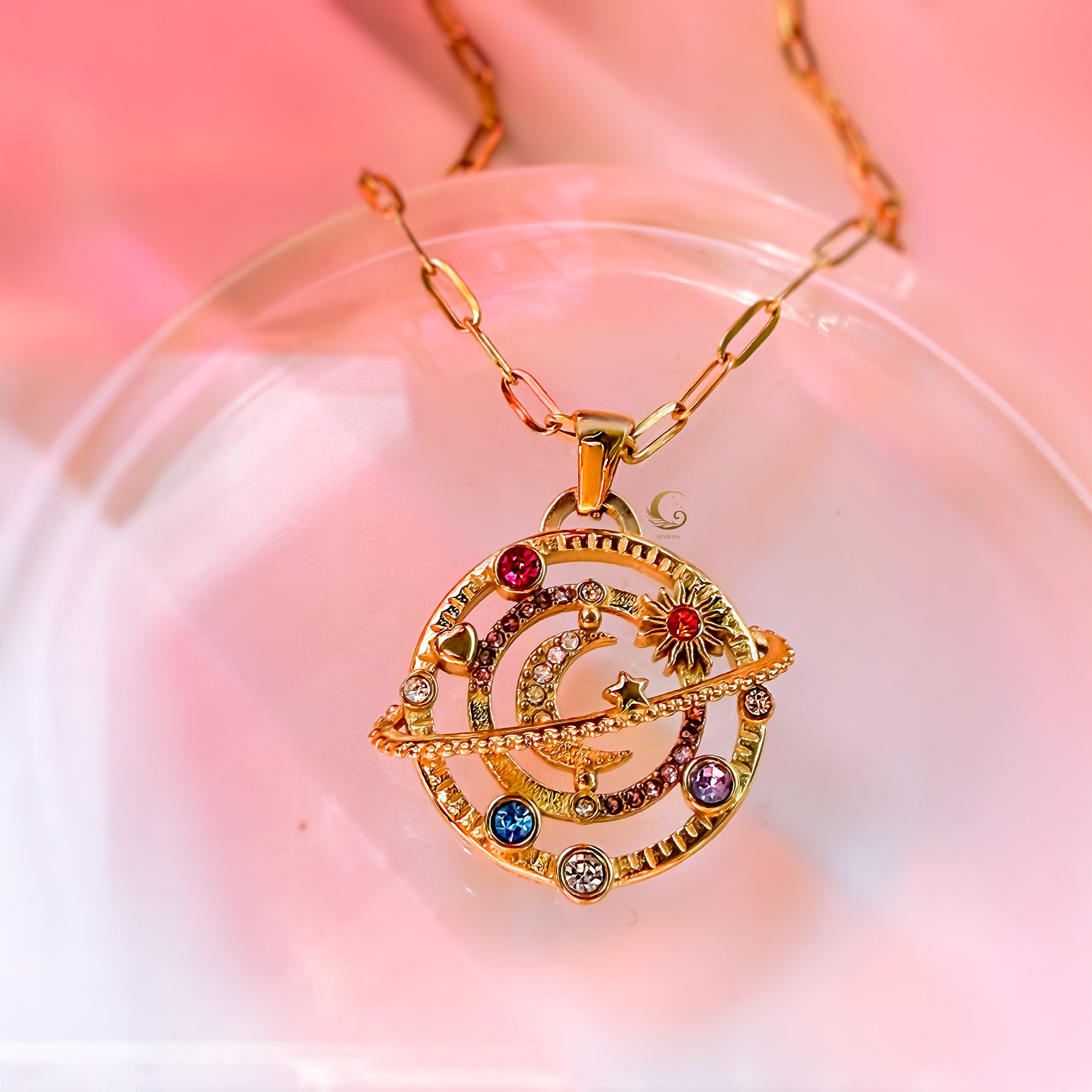 Gold celestial pendant with colorful stones and crescent moon on blush stone-like surface, AVYRONA jewelry.