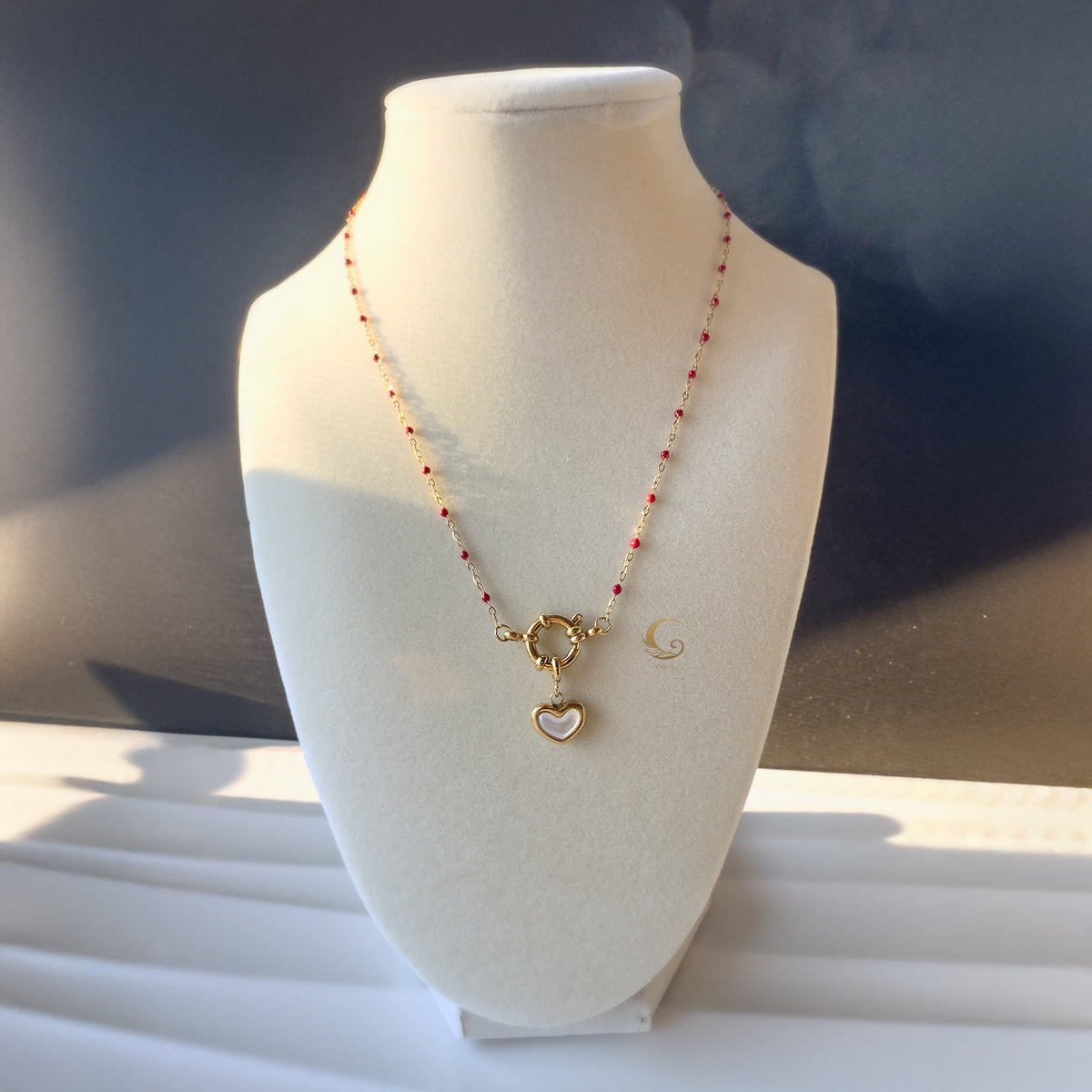 Rhea necklace with red beads and heart charm shown on white display bust in natural light