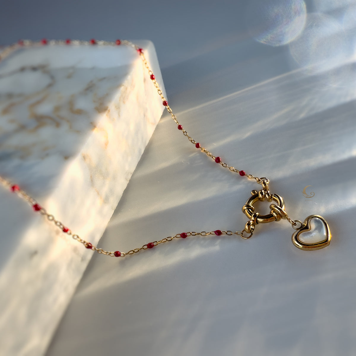 Delicate red beaded necklace with gold toggle clasp and mother-of-pearl heart pendant placed near marble slab