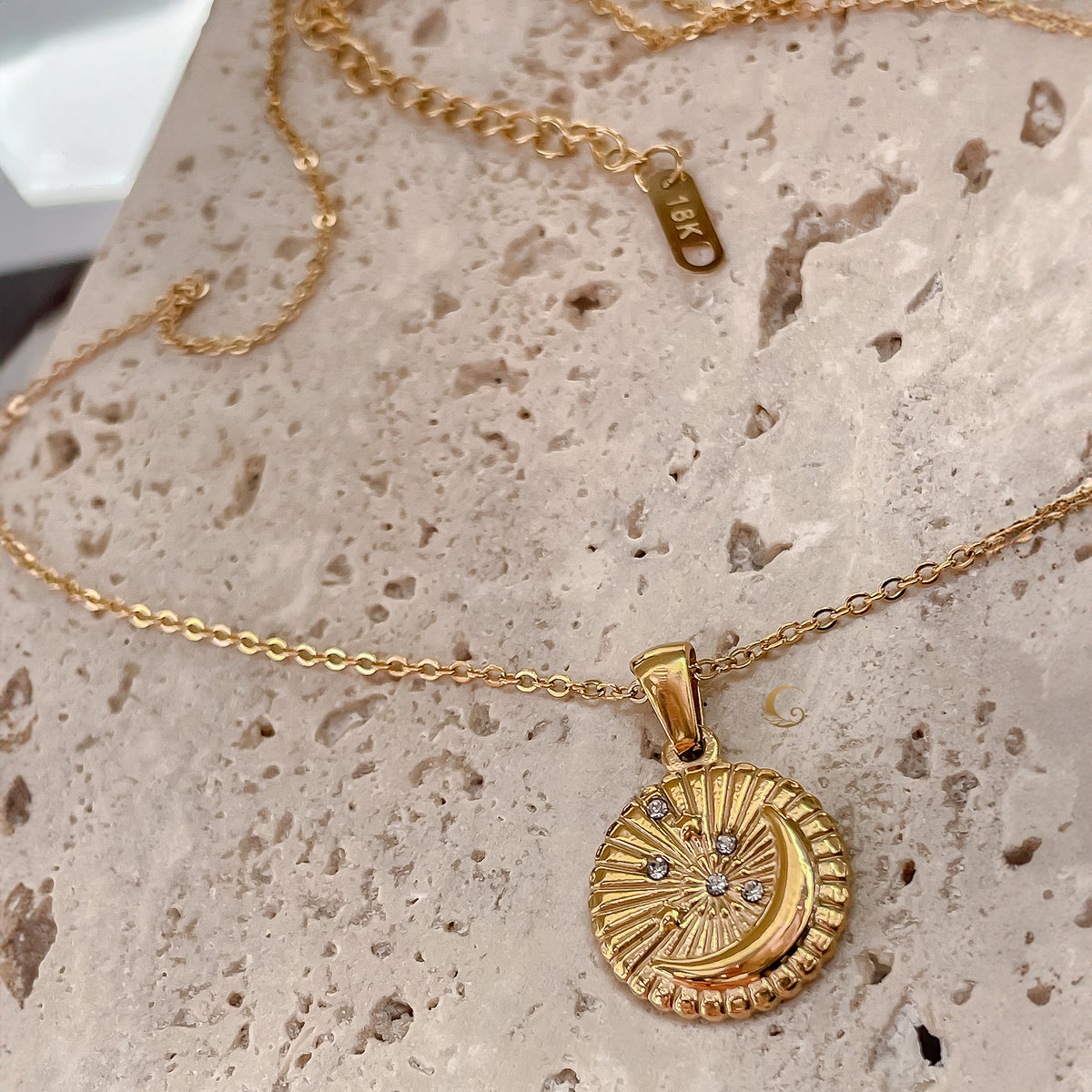 Selene Crescent Moon Necklace photographed on a natural stone surface, showing the gold-plated chain, crescent pendant with zirconia stars, and engraved 18K tag on the extension chain.