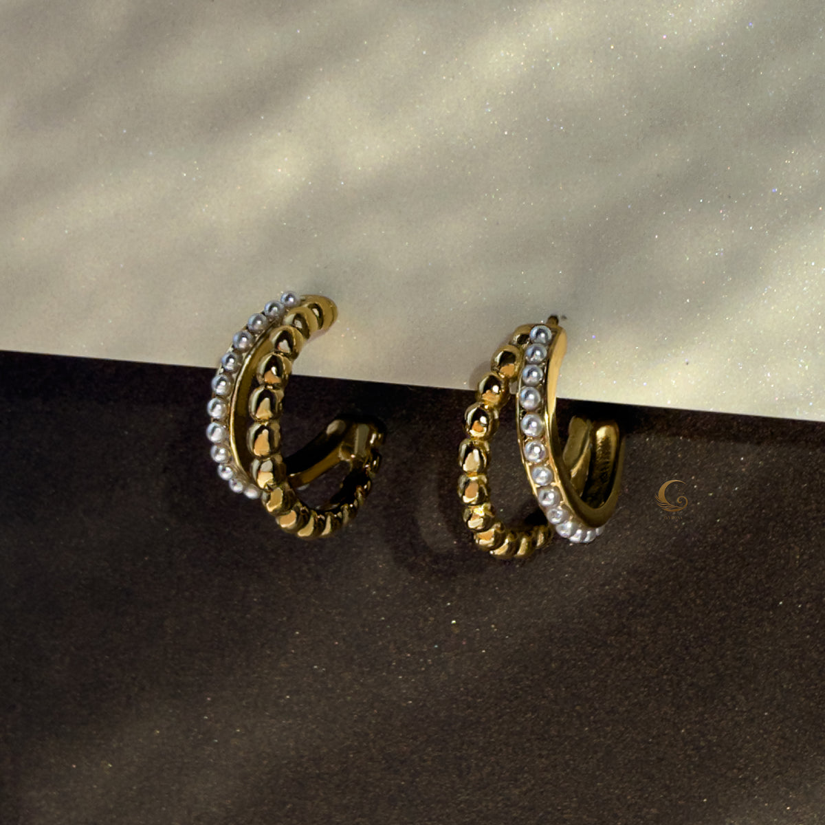 Serina dual gold hoop earrings glowing on deep dark surface with soft glimmer