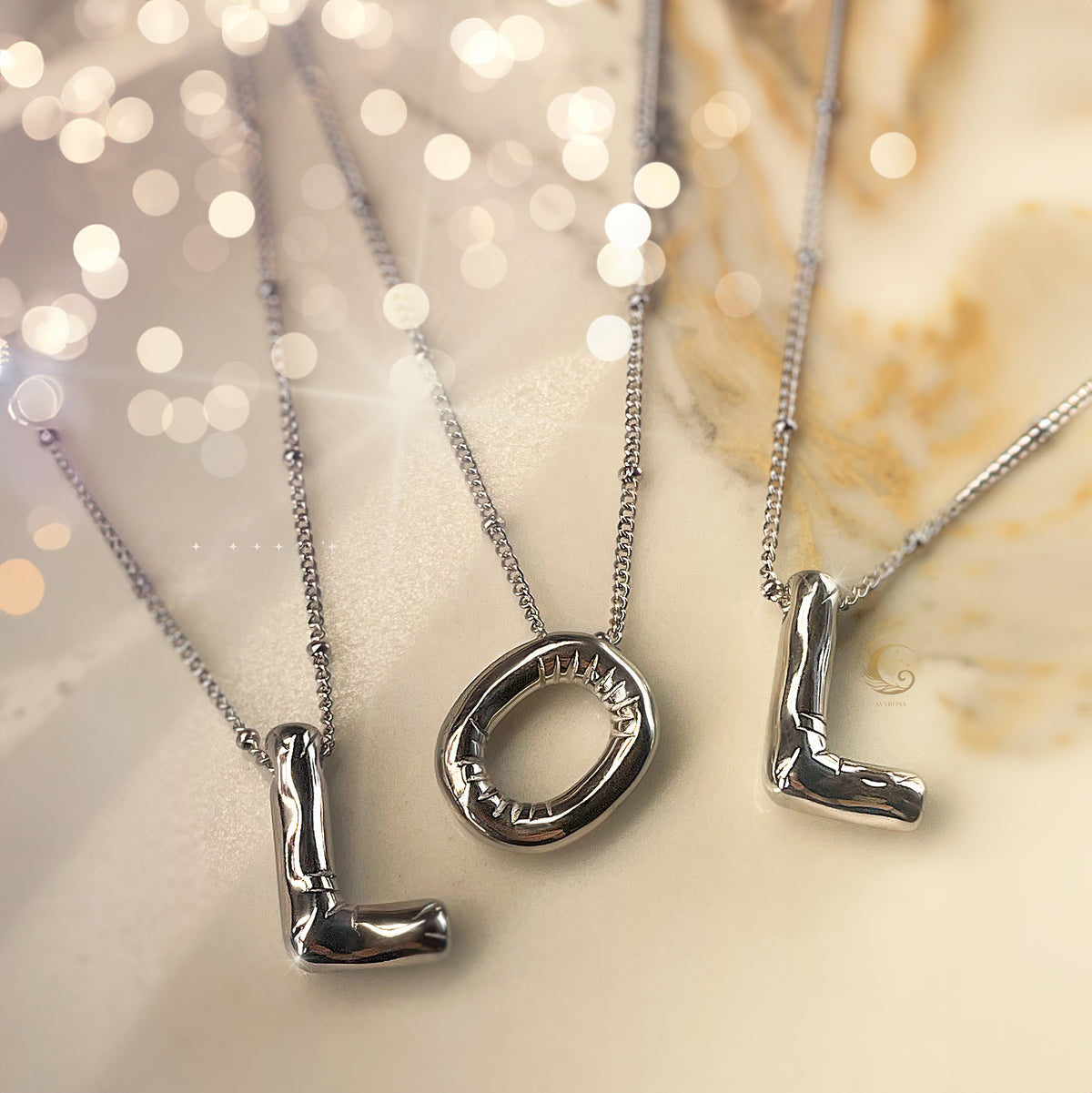 Three silver balloon-style letter necklaces forming the word “LOL” arranged on a dreamy marble background.