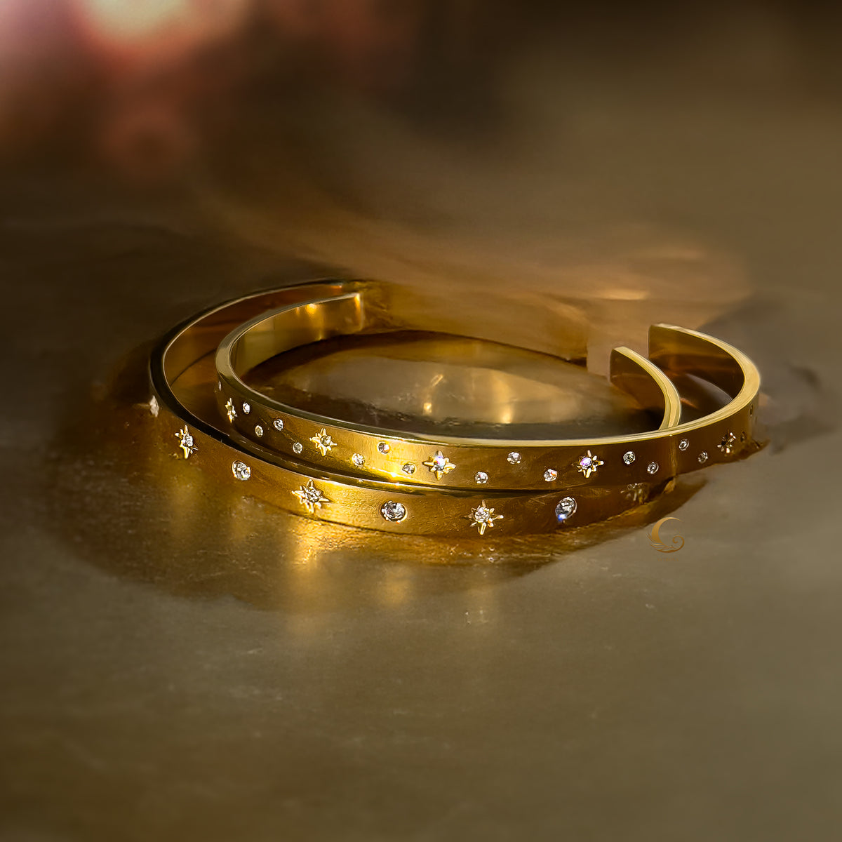 Two gold cuff bracelets with celestial star designs and cubic zirconia, resting on a golden satin surface.