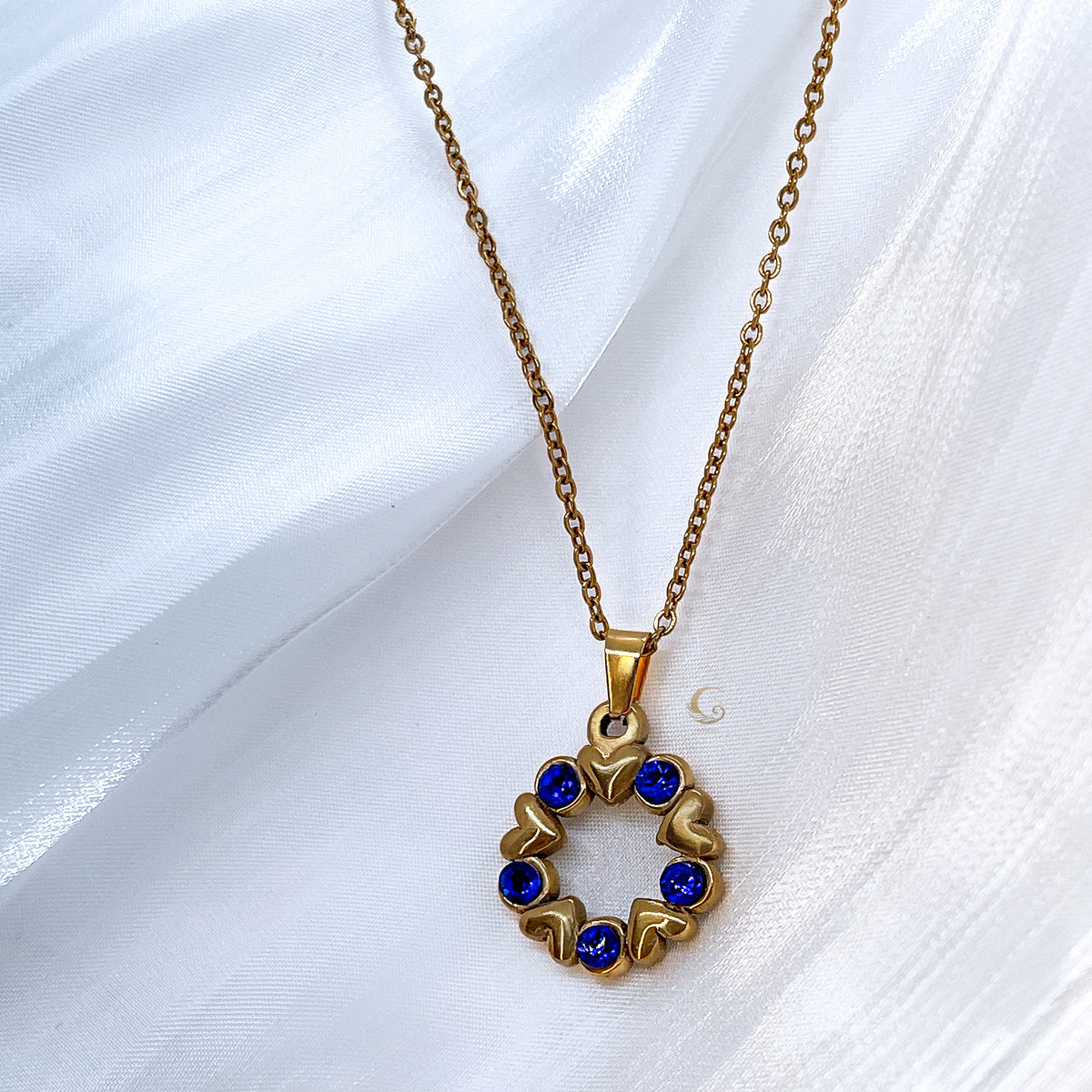 Close-up of Vega necklace with navy crystals and gold heart accents, placed on silky white fabric