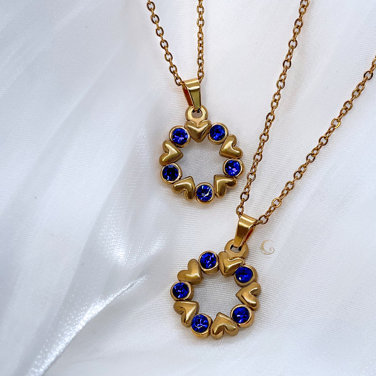 Two gold necklaces with navy crystal wreath pendants made of tiny hearts, displayed on soft white fabric