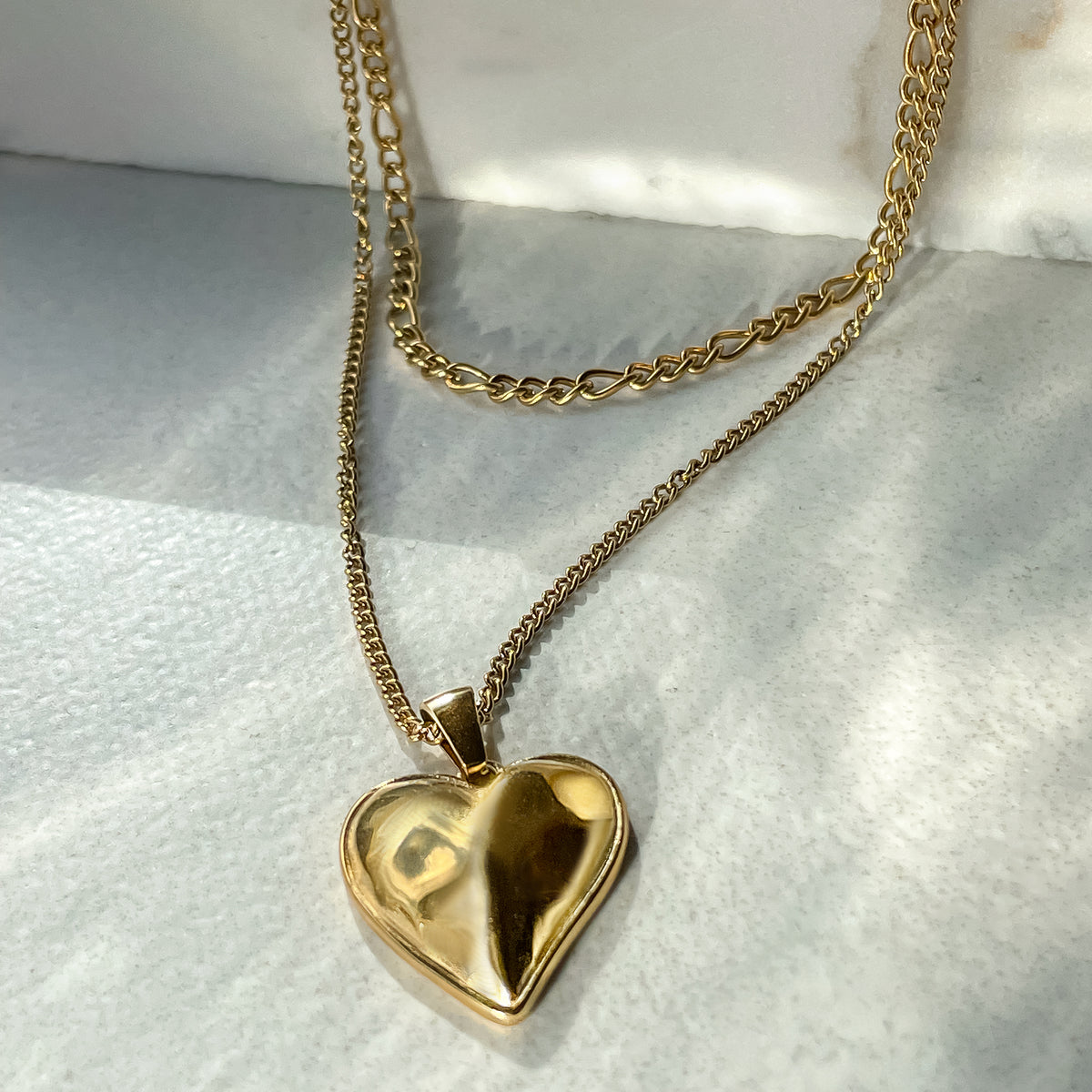 Close-up of layered gold heart necklace with reflective pendant and contrasting chains on soft white background.
